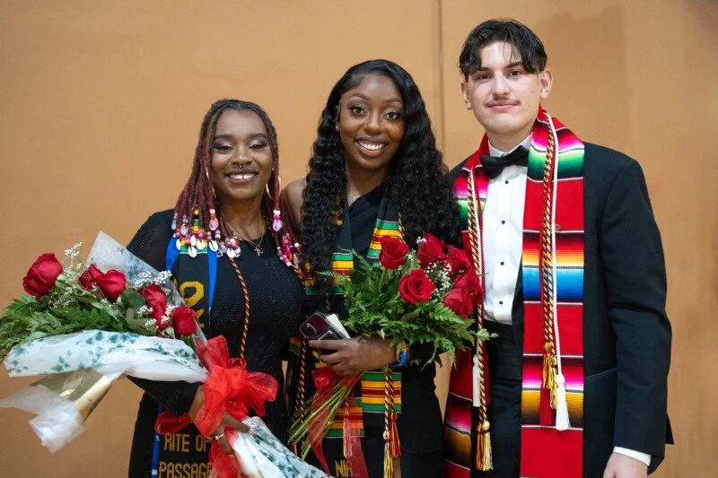 Three graduates standing and dressed in graduation attire.