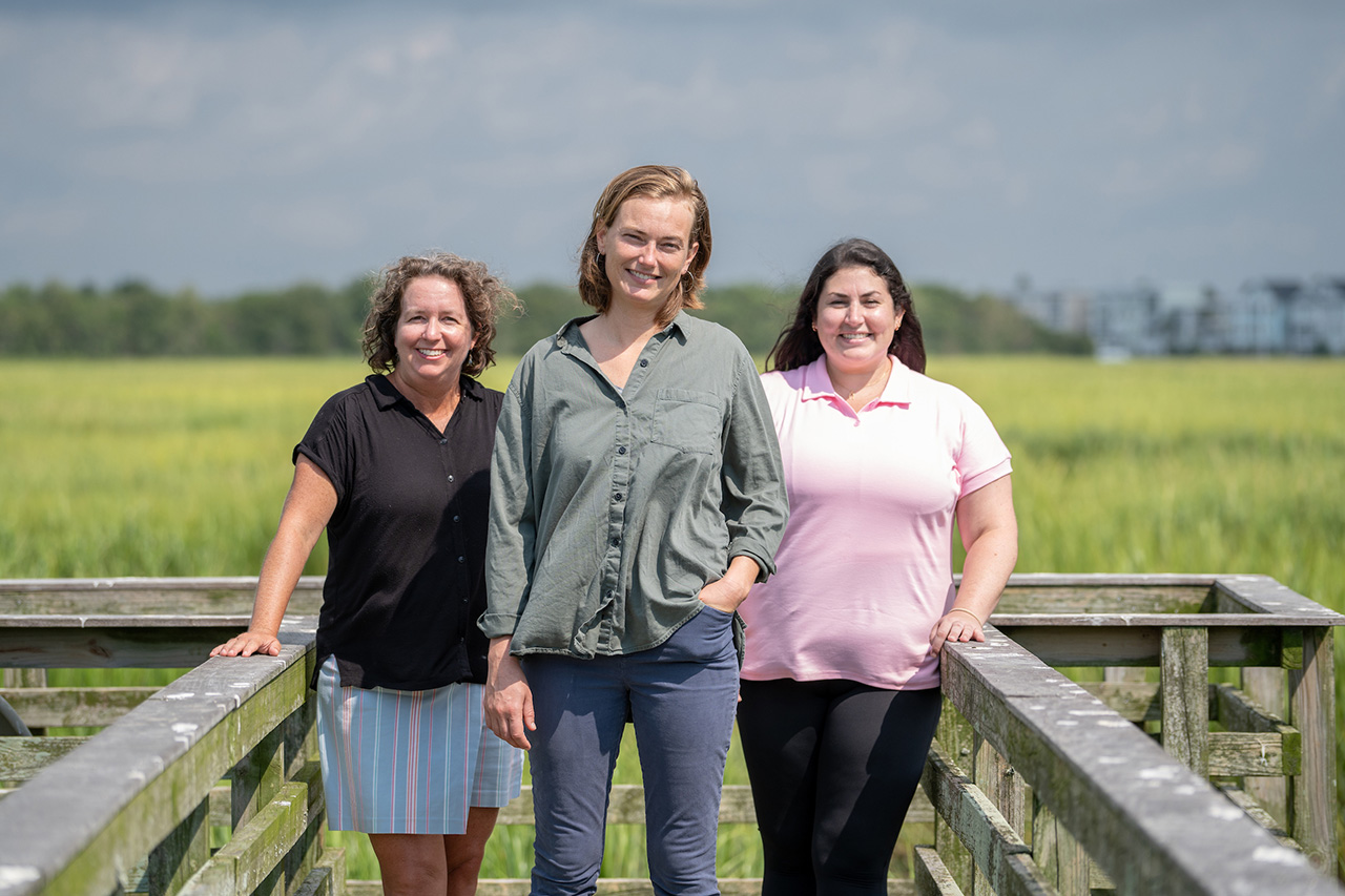 three women stand on wooden pier with marsh behind them and dark skies