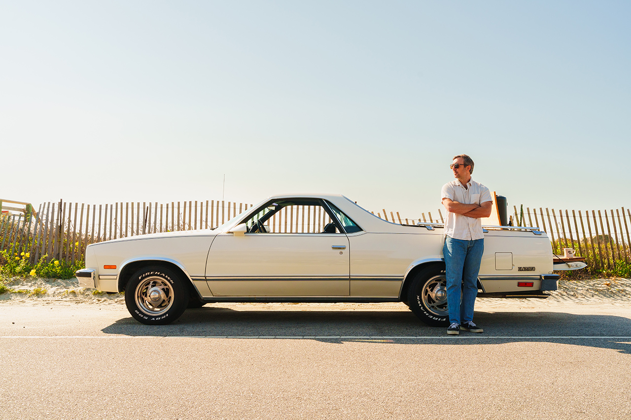man poses next to old car at the beach