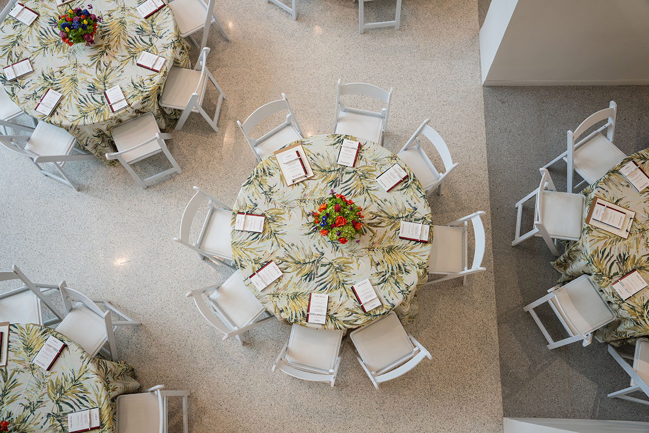 An overhead view of round tables with tropical-patterned tablecloths, white chairs, and floral centerpieces arranged in a spacious room.