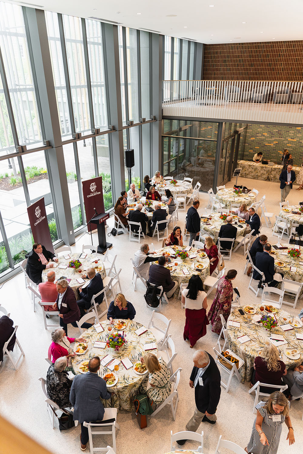 A group of people are gathered in a modern, well-lit atrium for a formal dinner event, seated at round tables with floral tablecloths and conversing with one another.