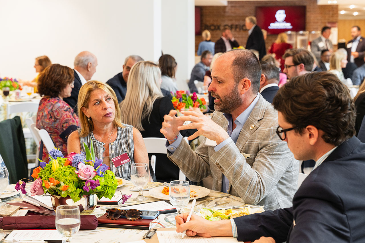 Attendees engage in conversation at a formal dining event, with colorful floral arrangements and business cards on the tables.