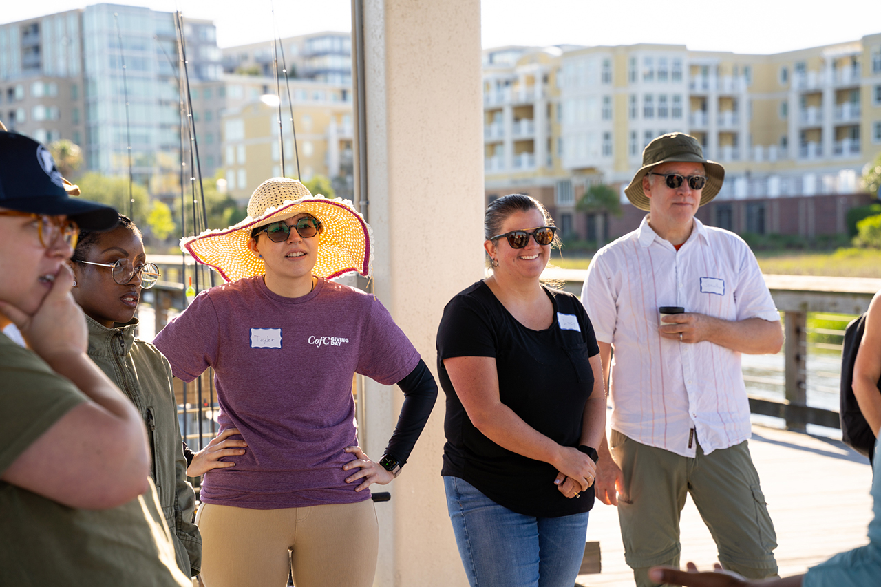 A group of people wearing hats and sunglasses stand on a dock, listening to someone speak, with modern buildings and water in the background.
