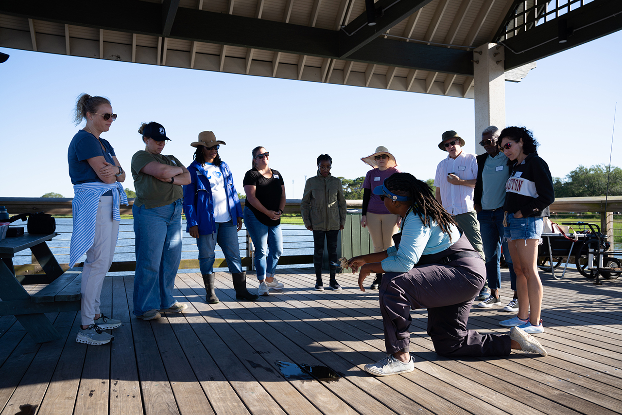 A group gathers on a wooden deck under a shelter as one person kneels and demonstrates something to the attentive audience.