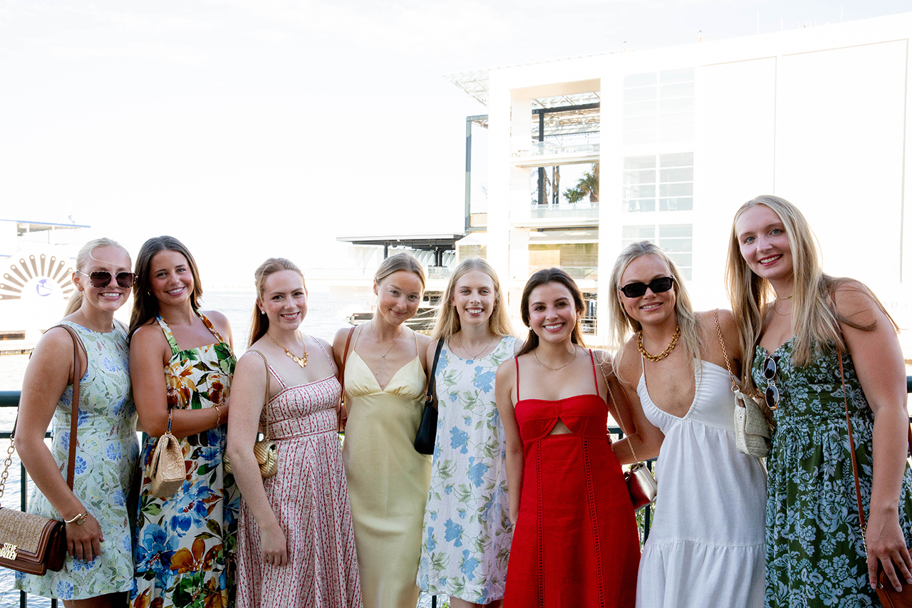 A group of eight individuals dressed in summer attire pose together outdoors near a waterfront, smiling for the camera.