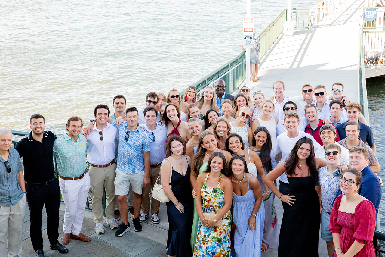 A diverse group of people poses together on a waterfront pier, smiling for the camera with the water and a railing visible behind them.