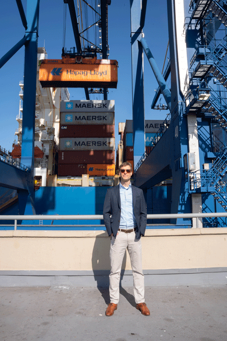 A person stands on a dock in front of large shipping containers and a crane with "Hapag-Lloyd" branding.