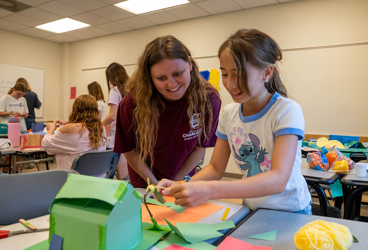 Two individuals collaborate on a craft project at a table, surrounded by colorful paper and art supplies in a classroom setting.
