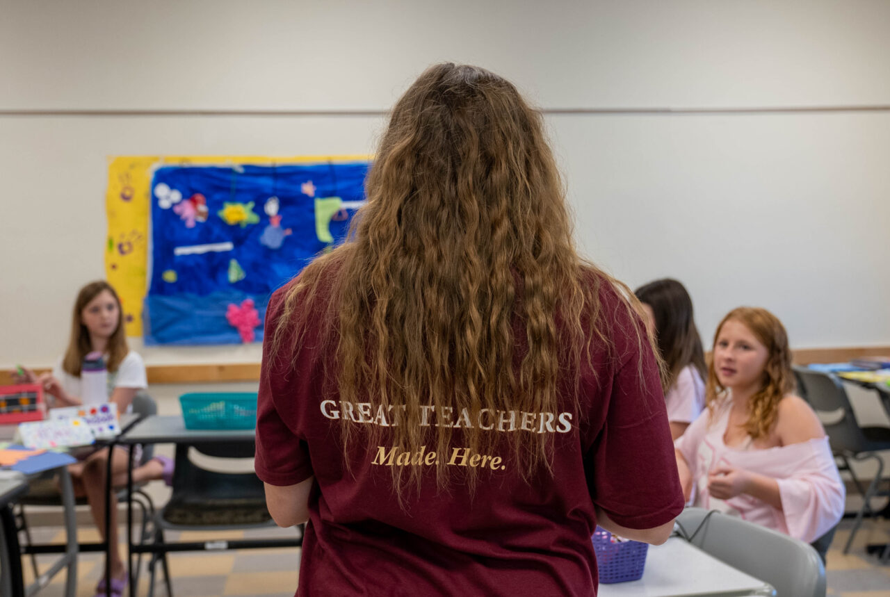 A person with long curly hair, wearing a maroon shirt that reads "GREAT TEACHERS Made Here," stands facing a group of children seated at tables in a classroom setting.