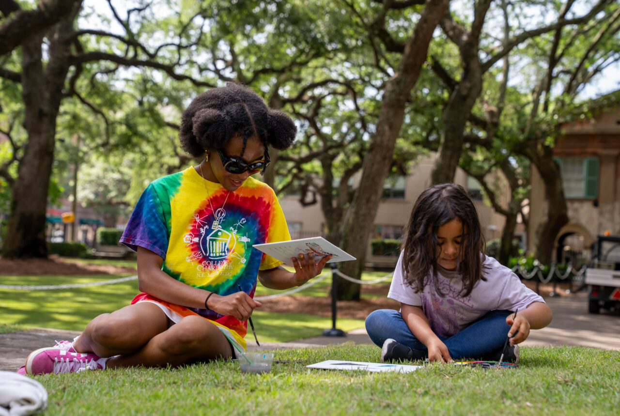 Two individuals sit on grass under trees, engaged in painting on paper, with one holding a tablet.