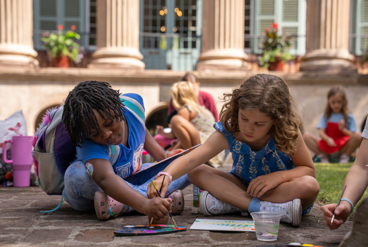 Two children sit on a stone surface outdoors, painting together with brushes and watercolors in front of a classical building.