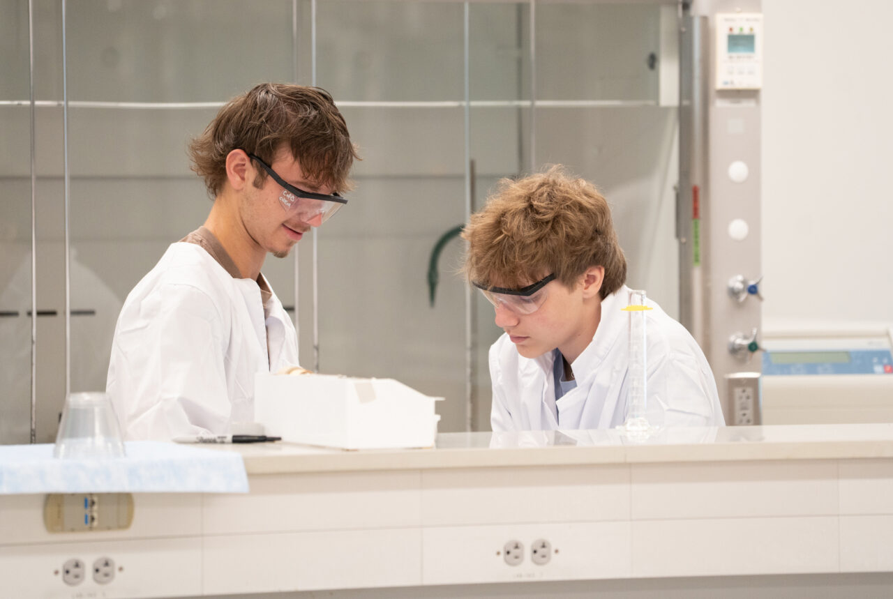 Two male students work together in lab wearing protective eye gear and lab coats