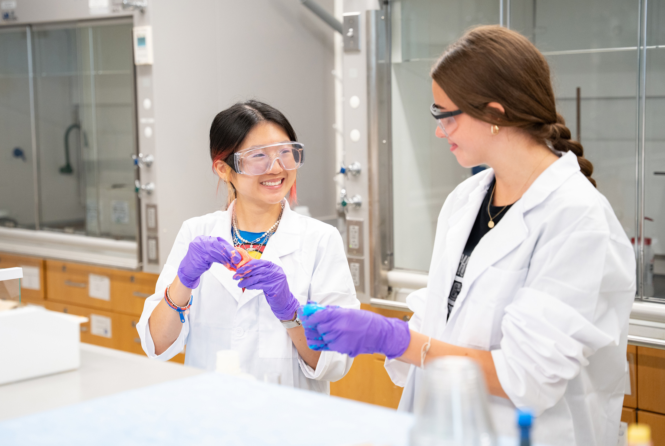 Two students smile in lab coats as they pull apart colorful material with purple gloves