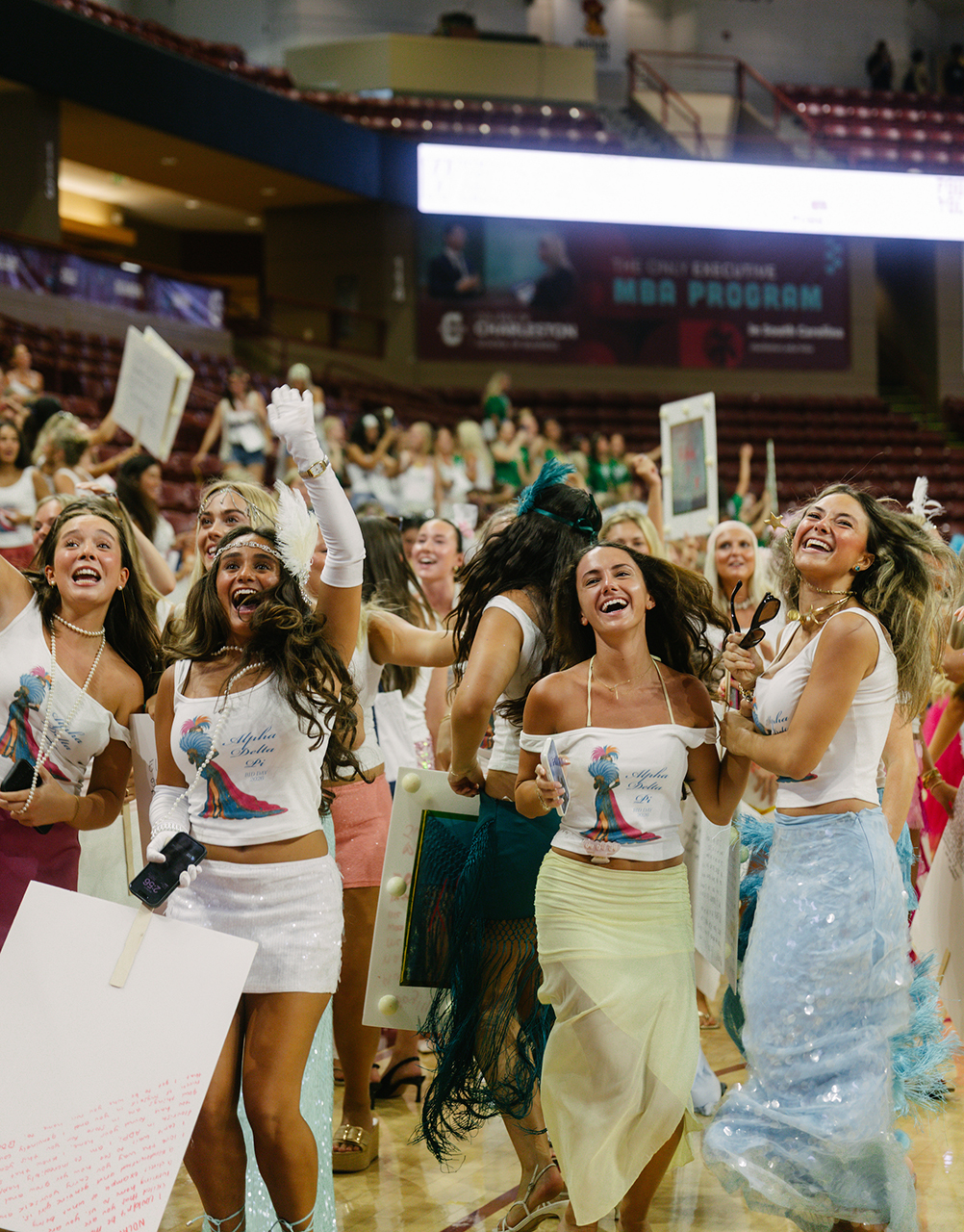 A group of smiling individuals in matching white tops and colorful skirts cheerfully hold signs and dance on a basketball court during an event.