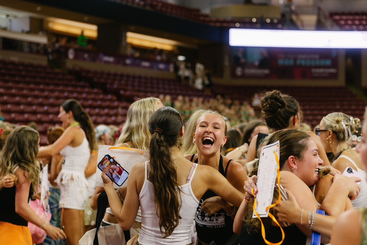 A group of young people are laughing and celebrating together in a large indoor arena, holding phones and papers, with empty red seats visible in the background.