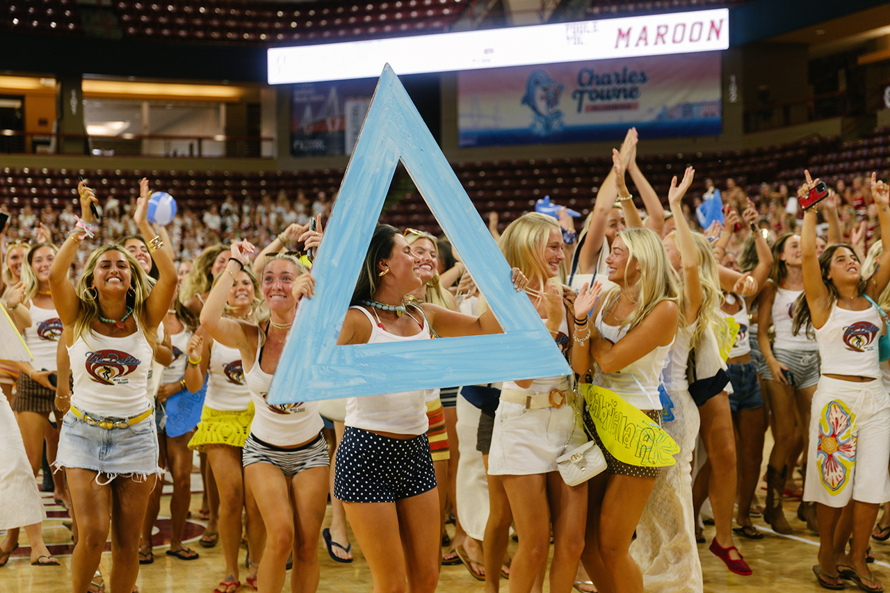 A group of young people in matching attire cheerfully hold up a large blue triangle sign at a crowded indoor event.