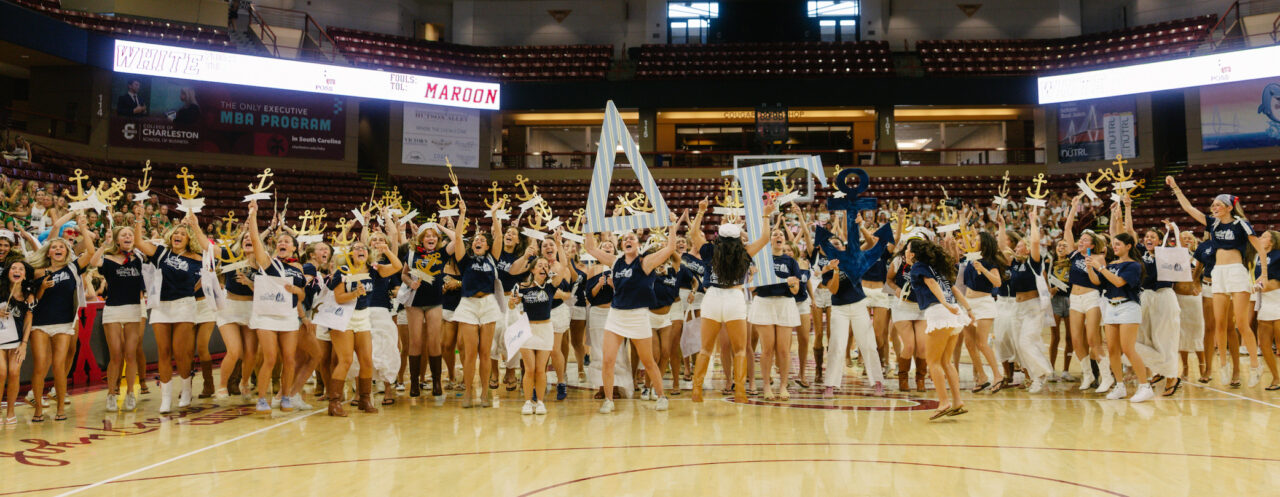 A group of cheerleaders in matching uniforms and gold headpieces celebrate on a basketball court, holding up large letters and trophies.