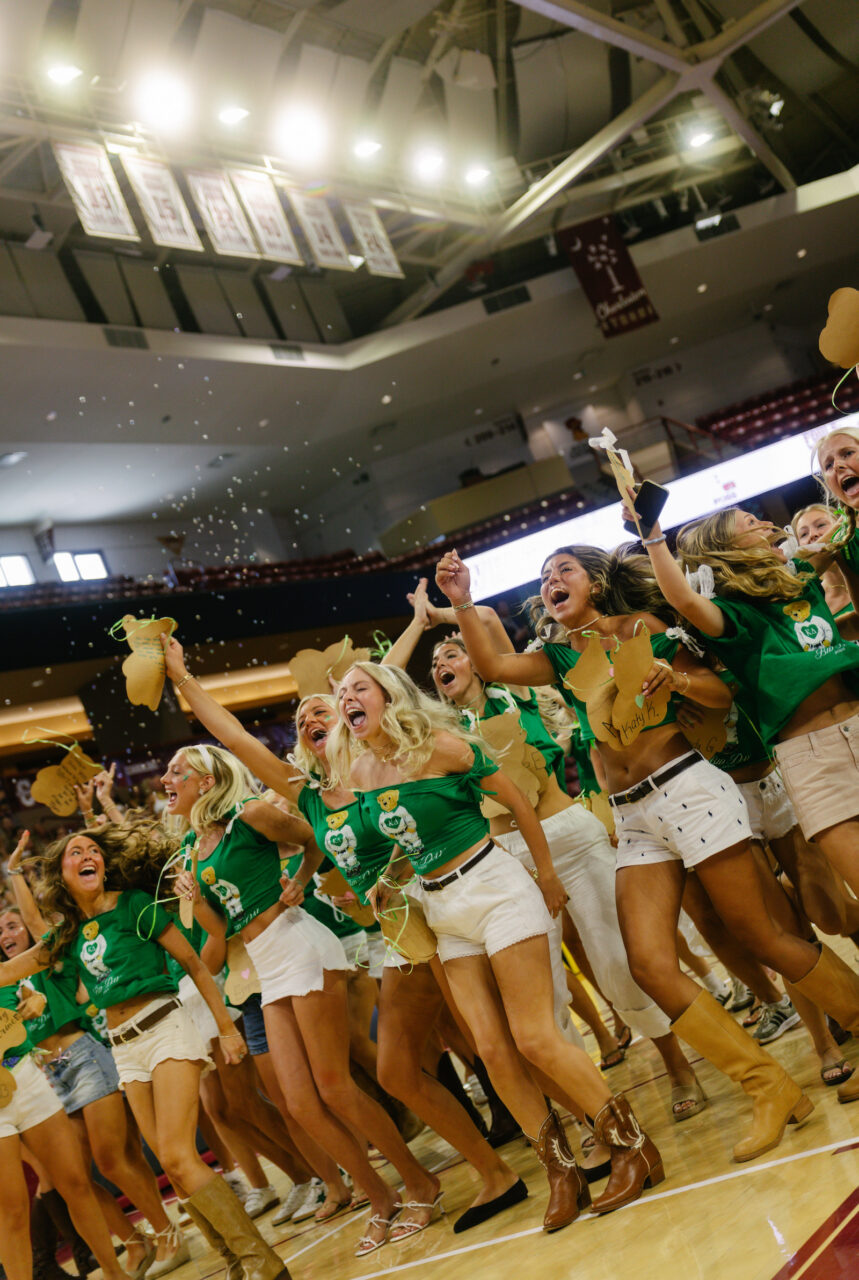 A group of cheerleaders in green tops and white shorts celebrate on a basketball court, throwing confetti and cheering enthusiastically.