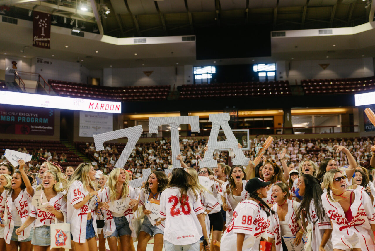 A group of enthusiastic supporters in white and red uniforms cheer with large cutout letters spelling "ZTA" at a sports event in a crowded arena.