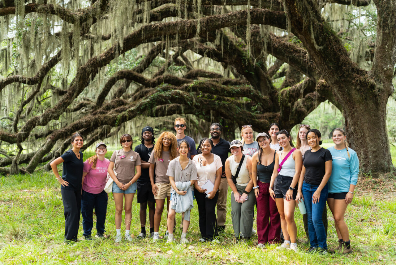 A diverse group of individuals poses together under a large, moss-draped tree in a grassy outdoor setting.