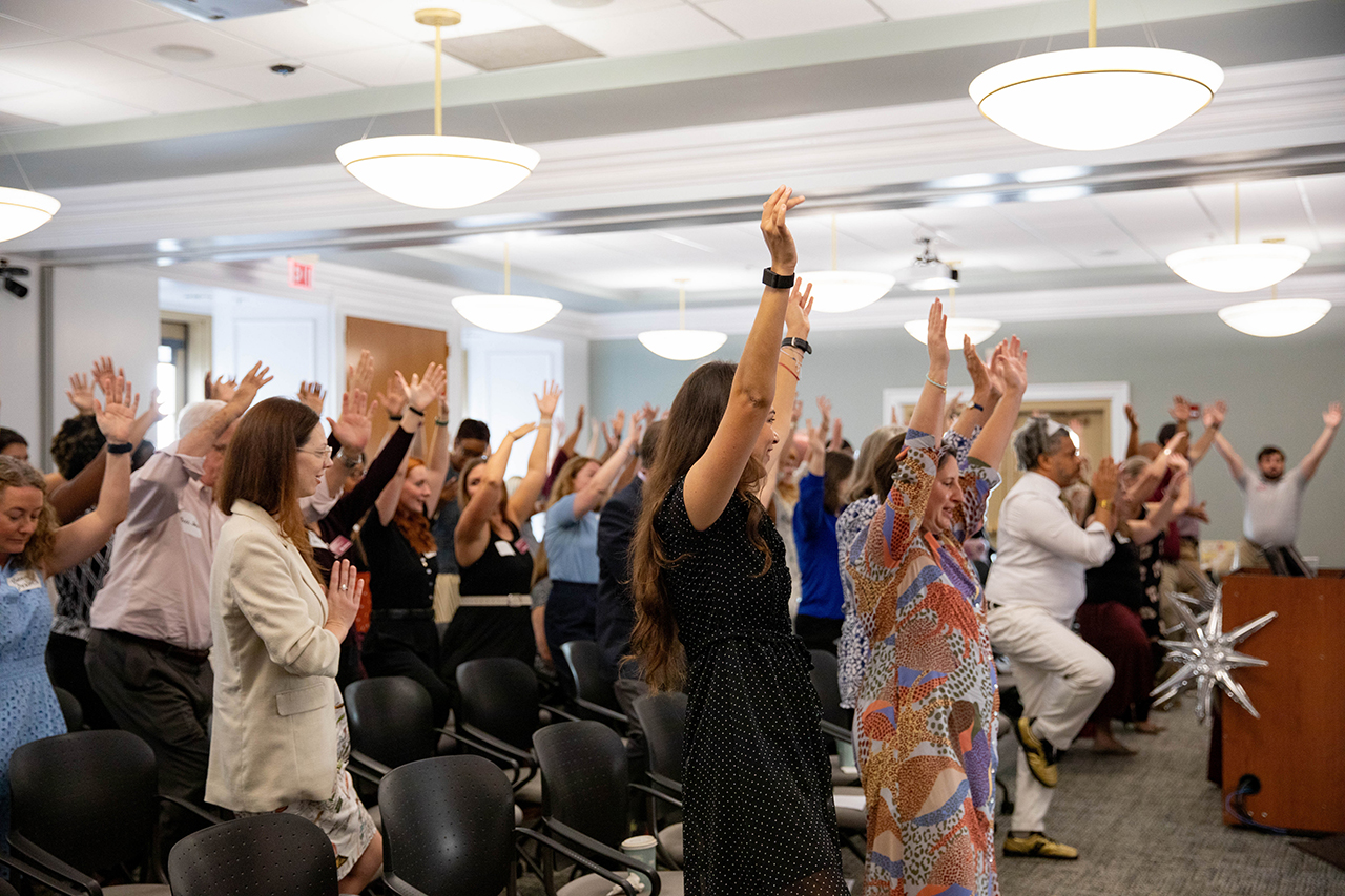 A diverse group of people in a conference room raise their hands in unison during a group activity.