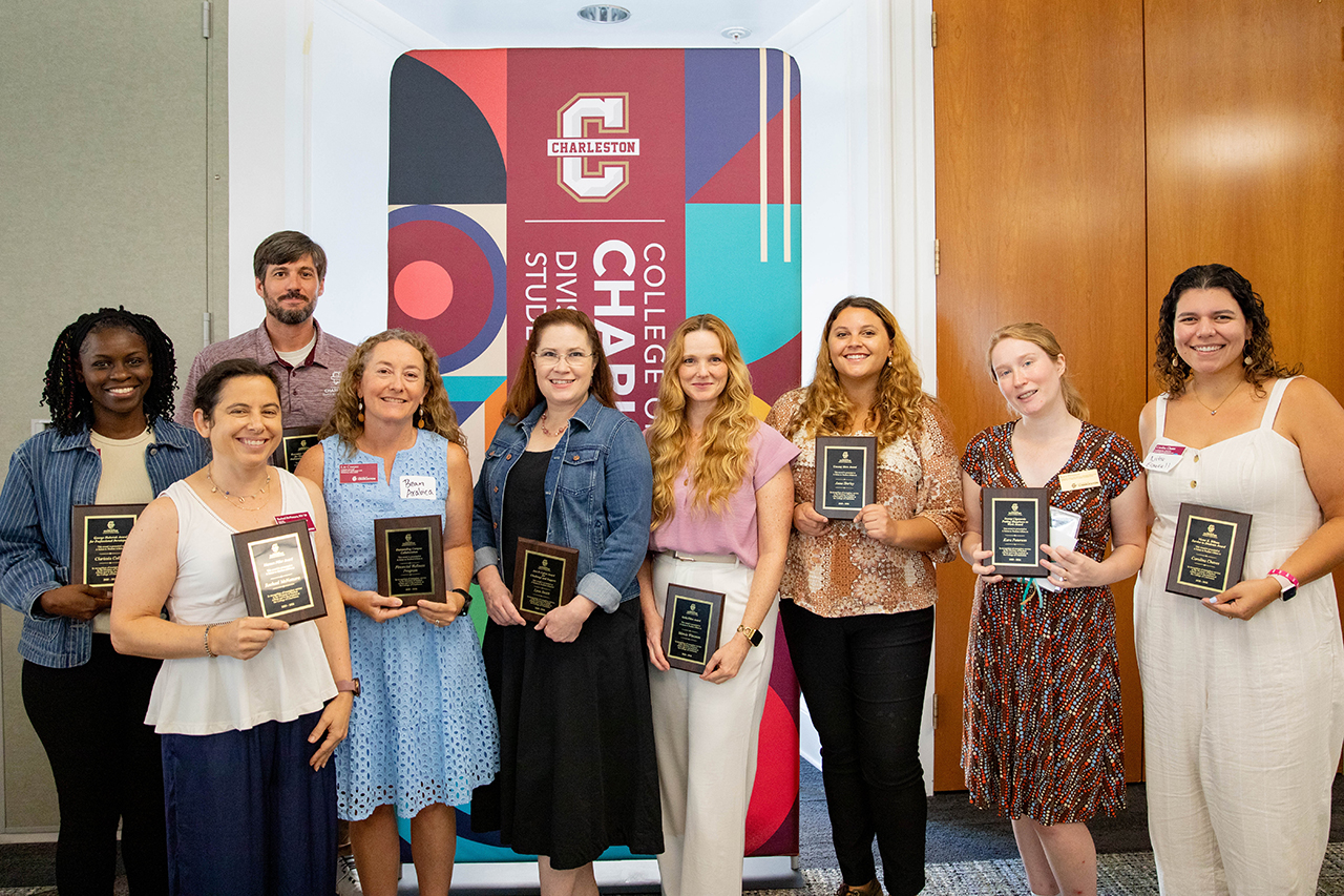 A group of eight individuals stand together indoors, each holding a framed award, in front of a colorful backdrop featuring the logo and name of Charleston College.