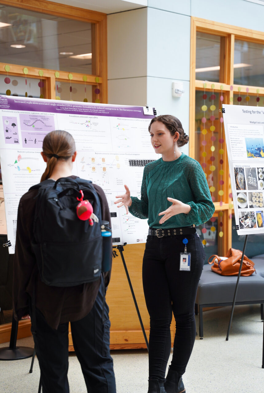 A person in a green top stands beside a scientific poster, gesturing while speaking to another individual with their back turned, in what appears to be an academic or research presentation setting.