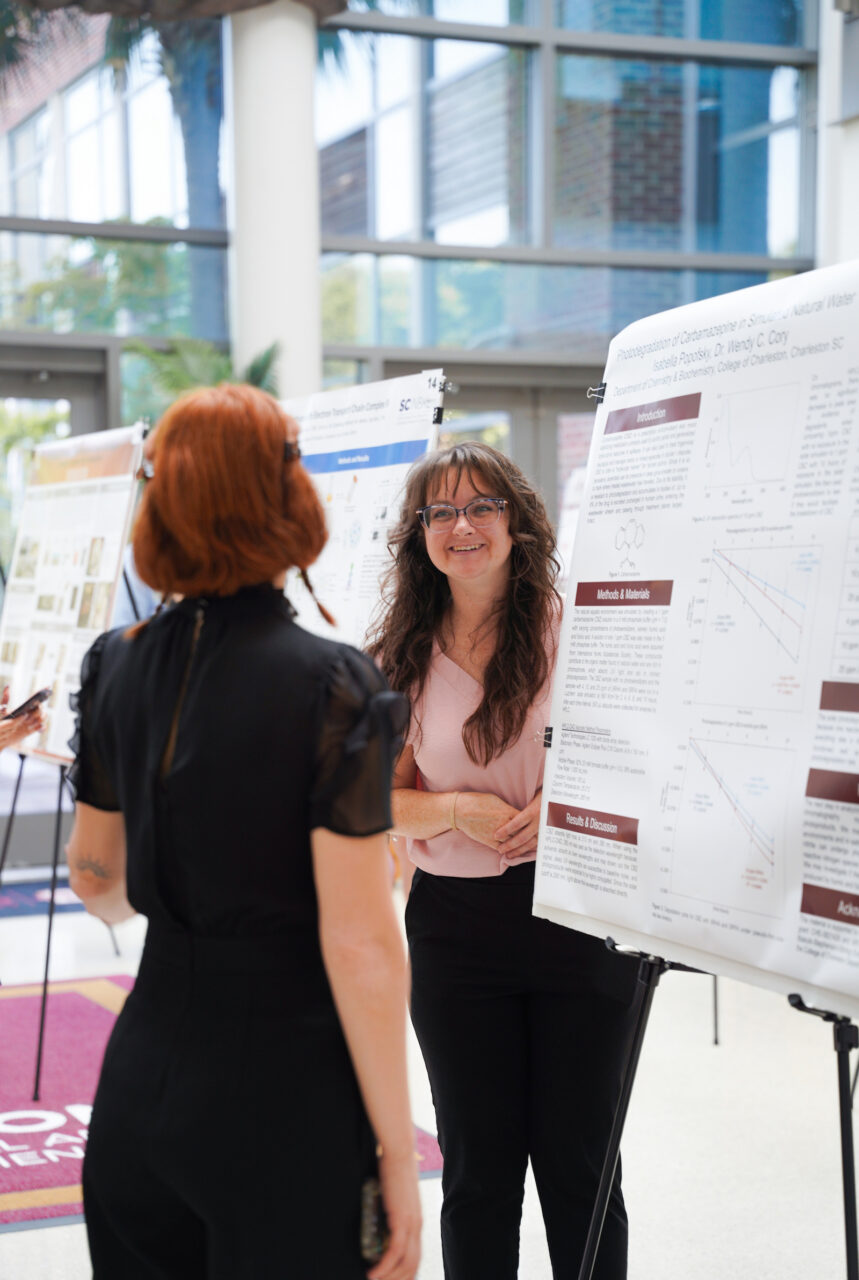 Two individuals engage in conversation at an academic conference, with one presenting research findings displayed on a poster board.