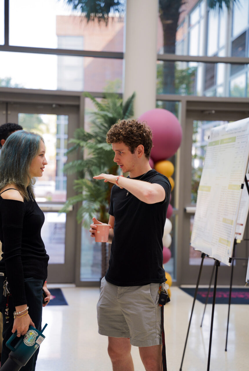Two individuals engage in conversation indoors, with one gesturing while holding a cup and the other listening, near a display board and colorful balloons.