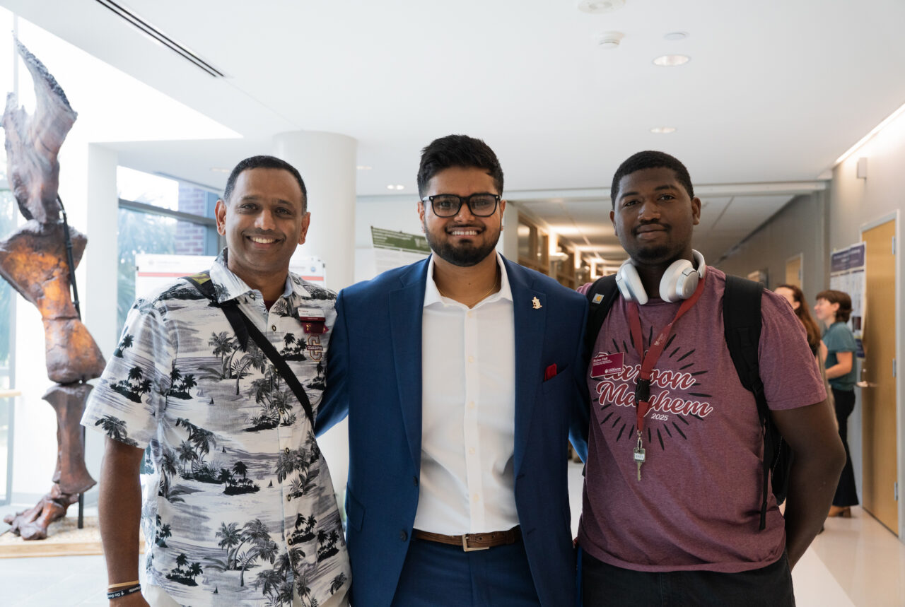 Three individuals pose together in a modern indoor space, with one wearing a tropical shirt, another in a suit, and the third in a casual t-shirt with headphones around his neck.