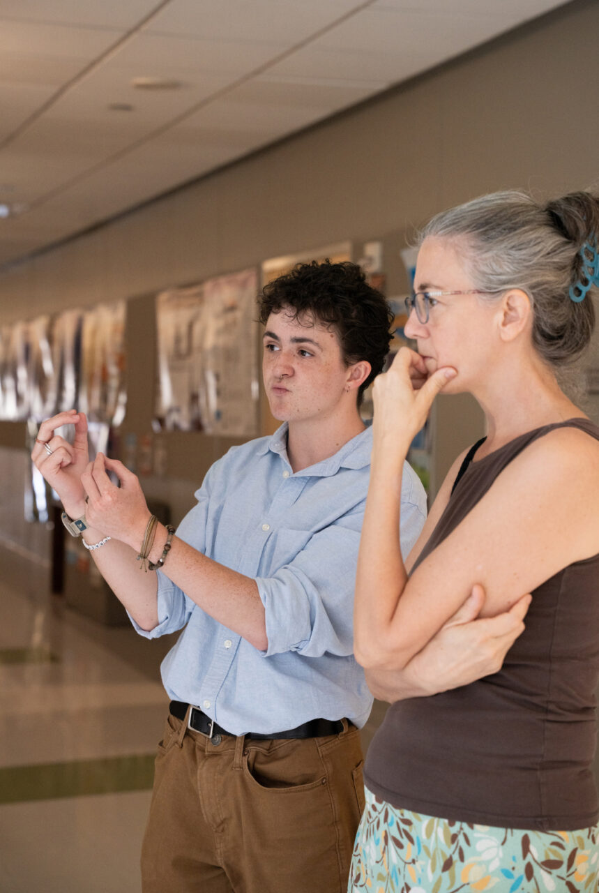 Two individuals stand in a hallway, one gesturing with hands while the other observes thoughtfully, both focused on something off-camera.