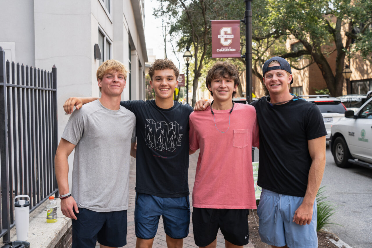 Four young people pose together on a sidewalk in front of a building with a Charleston University banner.