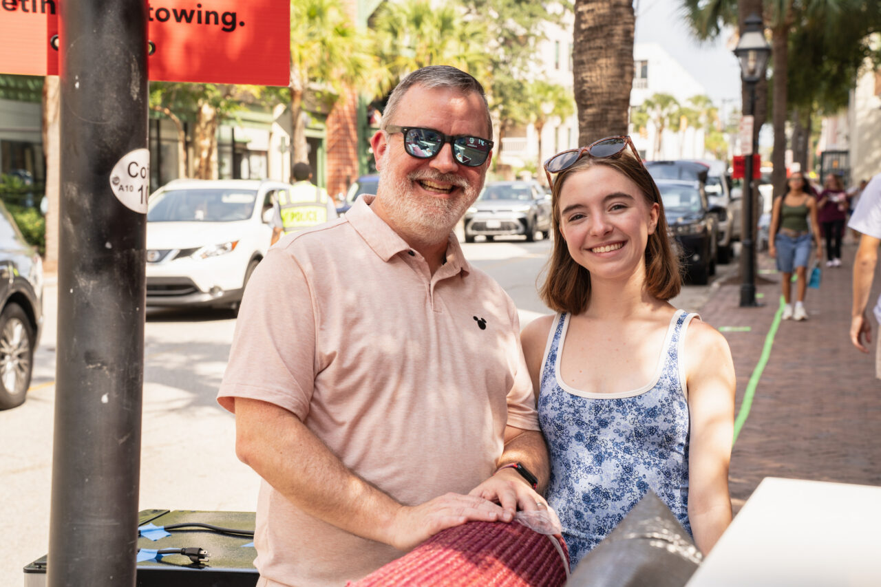 A smiling adult and a young person stand together on a sunny street, surrounded by palm trees and urban scenery.