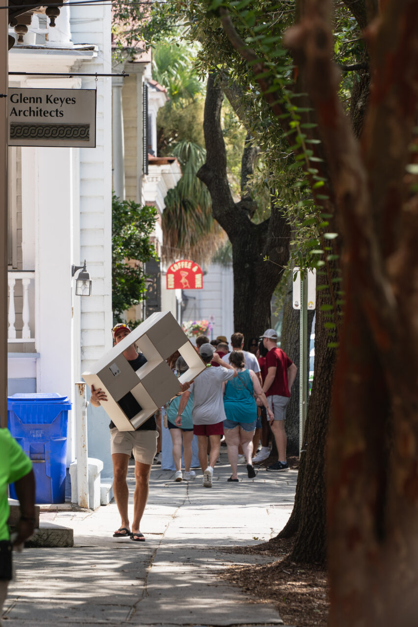 A person carries a large cardboard box down a tree-lined sidewalk while others walk nearby.