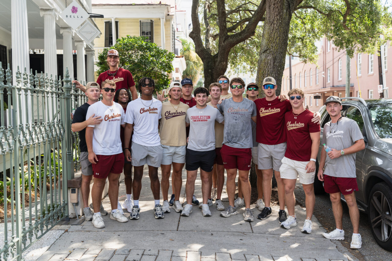 A group of young athletes in Charleston Athletics apparel pose together on a sidewalk near a historic building and tree.