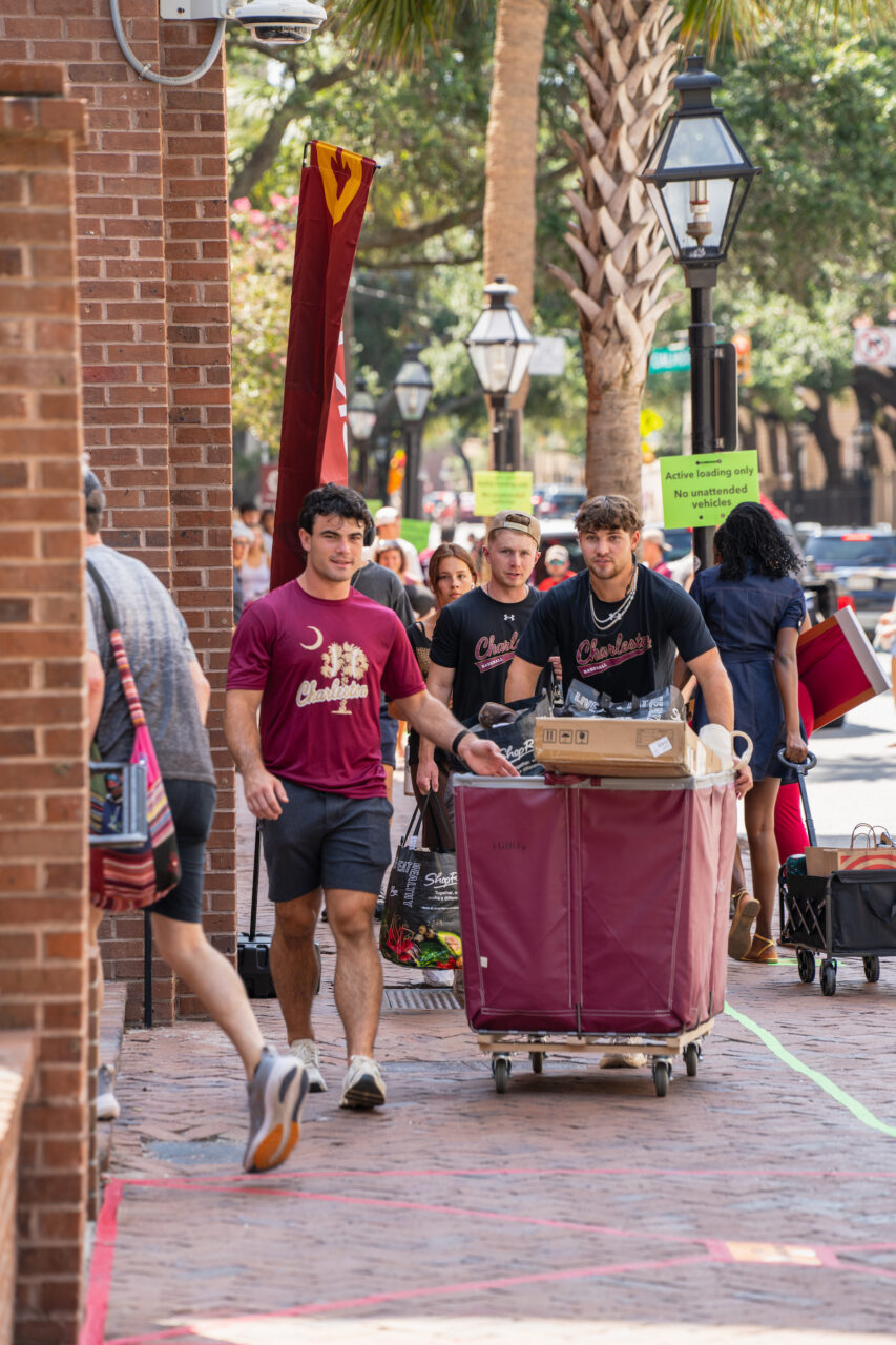 Several individuals walk along a brick-paved street, with one person pushing a cart loaded with boxes and shopping bags, while others carry bags and wear shirts featuring “Charleston” branding.