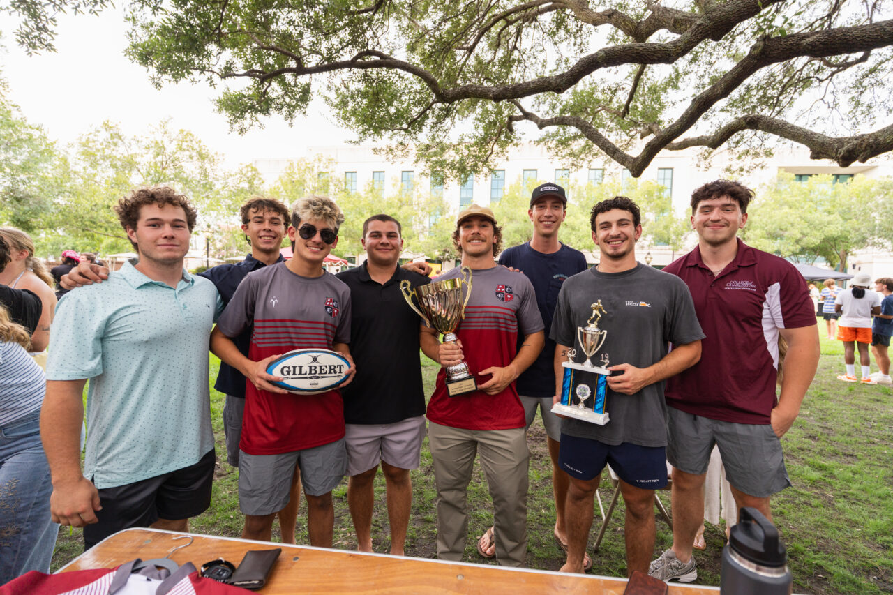 A group of young athletes stands outdoors under a large tree, proudly holding trophies and a rugby ball, celebrating their achievement.