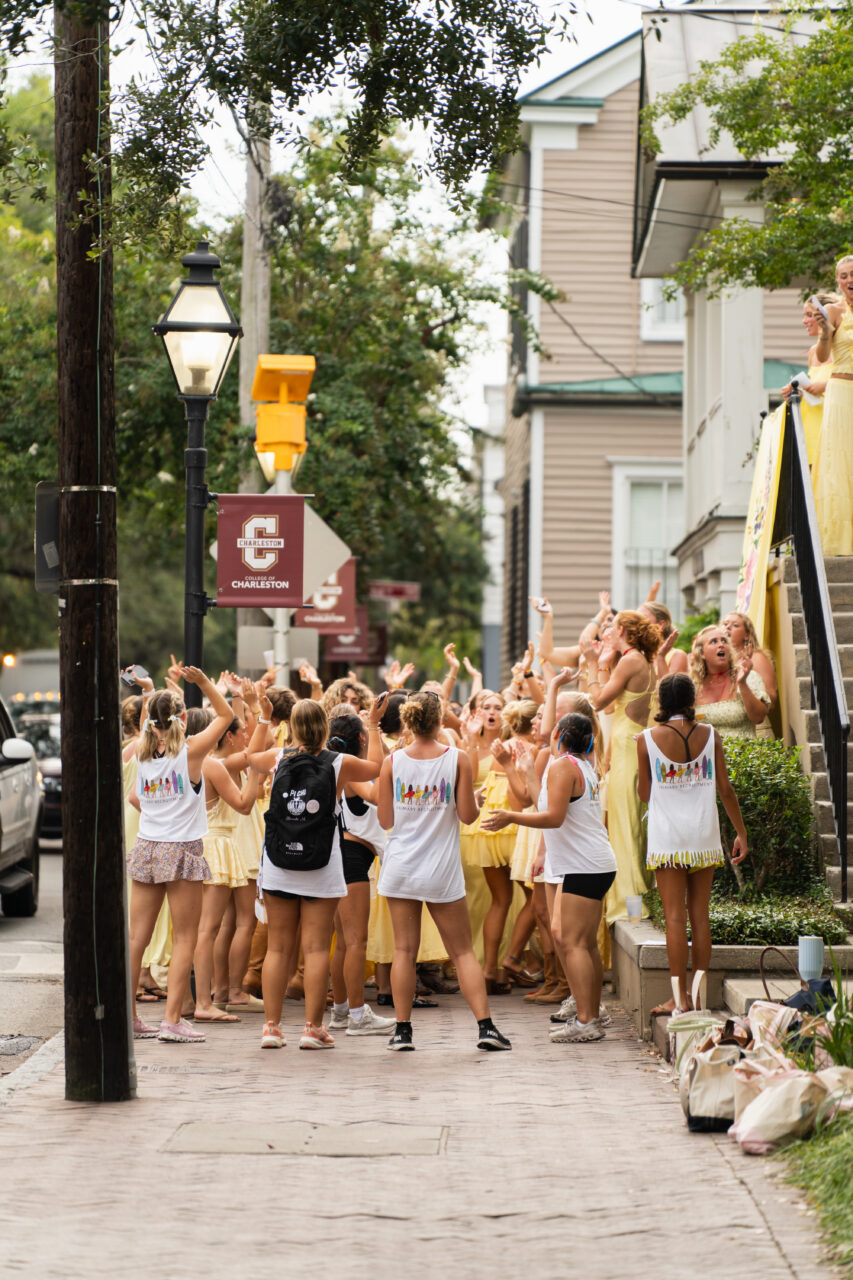 A group of people in yellow dresses and white shirts gather on a sidewalk, cheering and raising their arms, with some standing on steps beside a building.