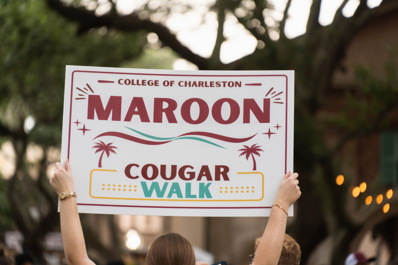 A person holds a sign for the College of Charleston’s Maroon Cougar Walk, featuring palm trees and festive graphics.