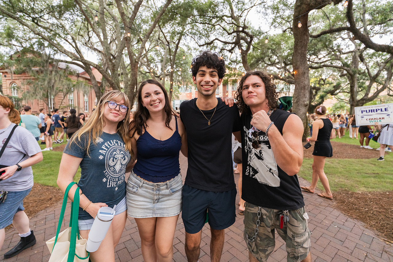 Four young people pose together on a brick path at an outdoor event, surrounded by trees and other attendees.
