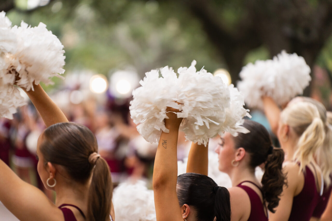 A group of cheerleaders in maroon uniforms raise white pom-poms high in unison outdoors.