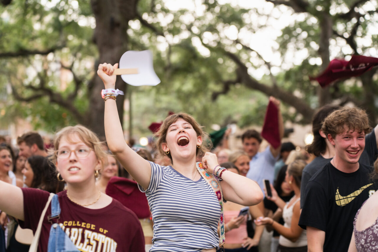 A joyful crowd at an outdoor event, with one person enthusiastically waving a white fan while others cheer and celebrate.