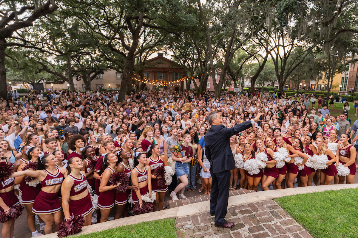 A person in formal attire stands on a stone platform, gesturing to a large crowd of students and cheerleaders gathered outdoors under trees.