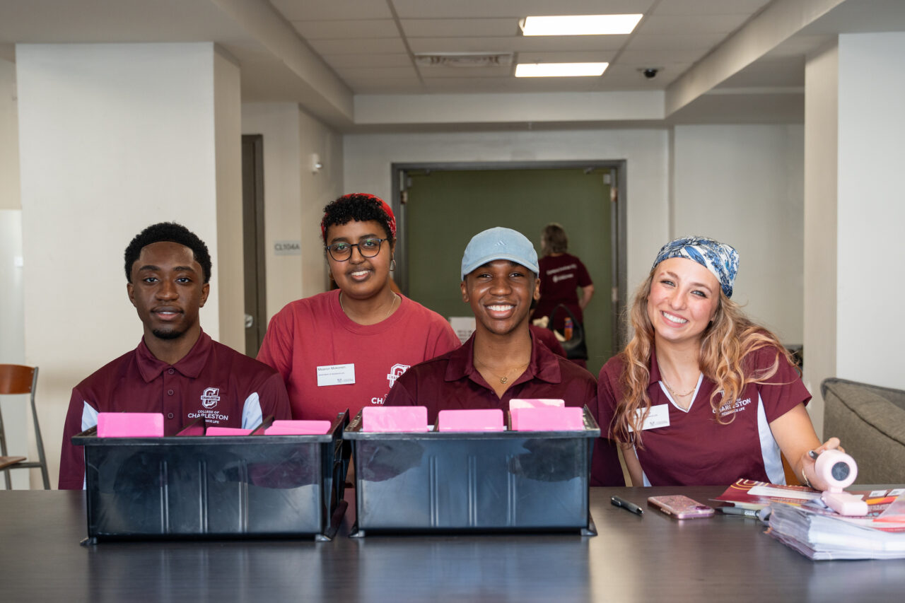 Four individuals in maroon shirts stand behind a table with pink folders, smiling at the camera in an indoor setting.