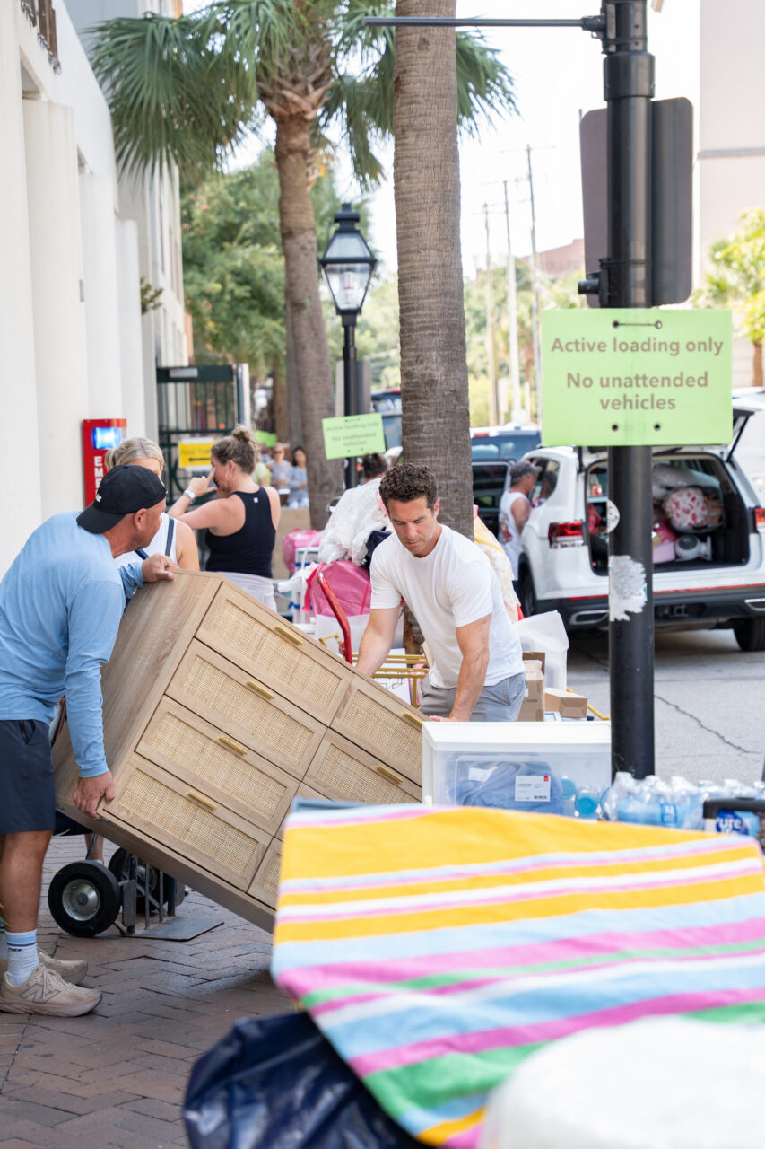 Two individuals are maneuvering a large wooden dresser on a dolly along a paved street lined with palm trees, near a vehicle with its trunk open.