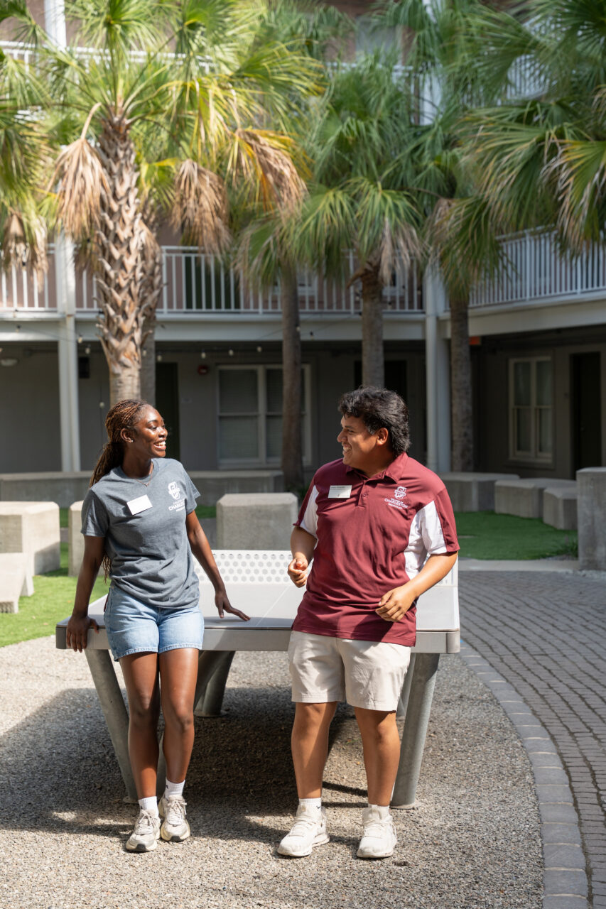 Two individuals in casual attire stand beside a bench outdoors, smiling and engaged in conversation under palm trees.
