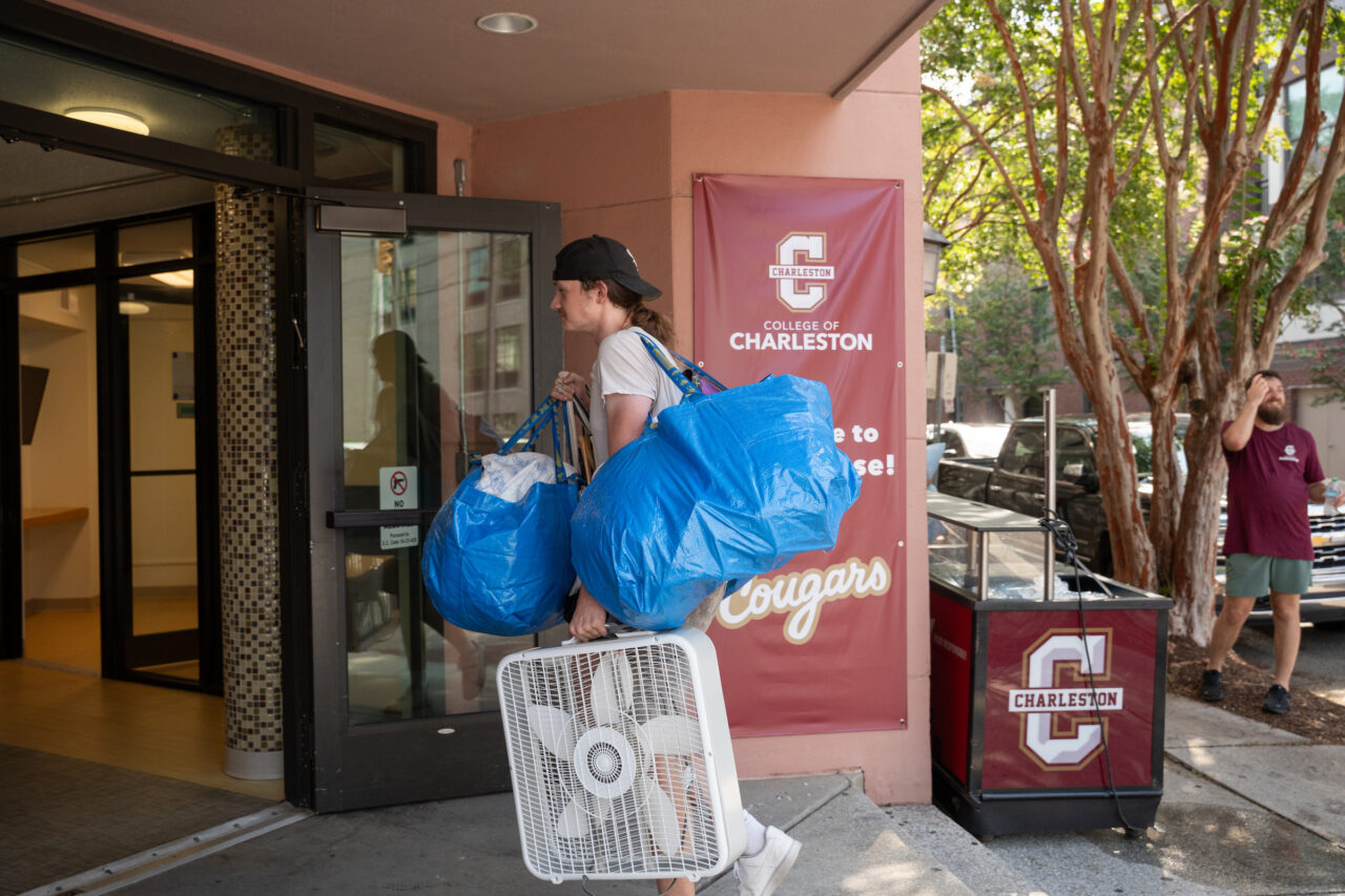 A person carries large blue bags and a white fan outside a building with a College of Charleston banner.