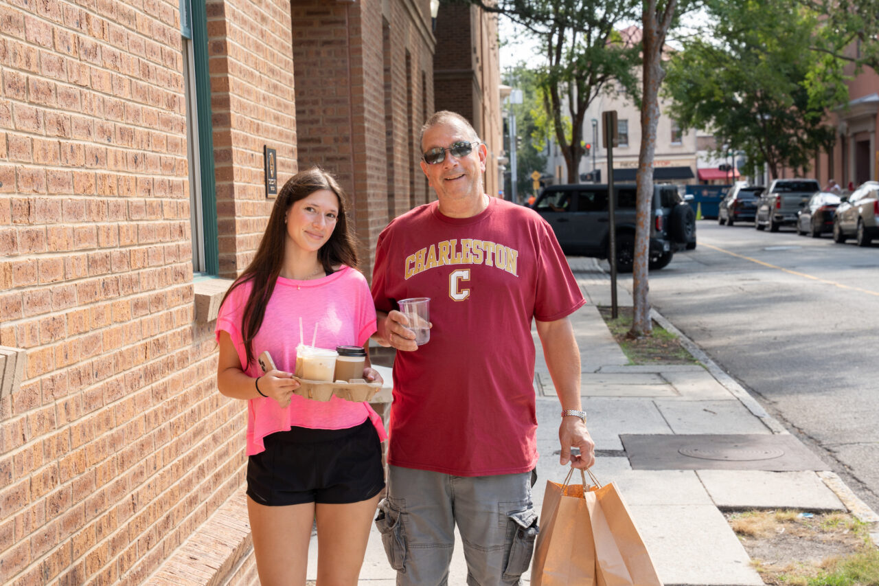 Two individuals walk along a sidewalk beside a brick building, each holding beverages and a shopping bag.