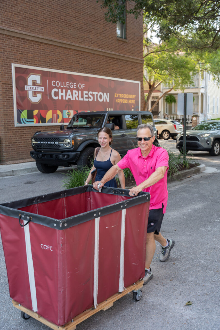 Two people push a large red cart with “COFC” branding down a street in front of a brick building with a banner for the College of Charleston.