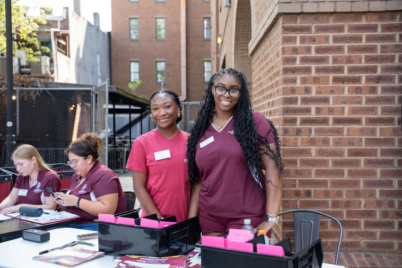 Two individuals in maroon uniforms stand smiling at an outdoor event, with others working at tables nearby.